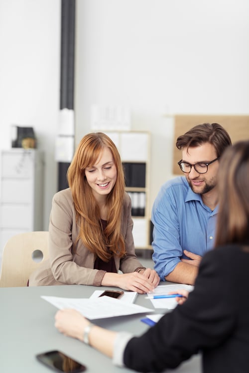 Young couple sitting in a meeting with an agent looking at a document together that she is presenting to them, view over the agents shoulder Young couple sitting in a meeting with an agent looking at a document together that she is presenting to them, view over the agents shoulder